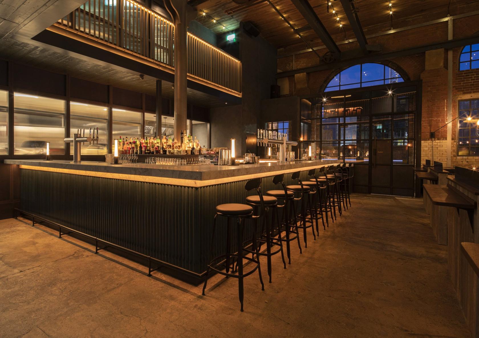 A restaurant with a long bar, high stools, exposed brick walls, and large arched windows at The Light Bar.