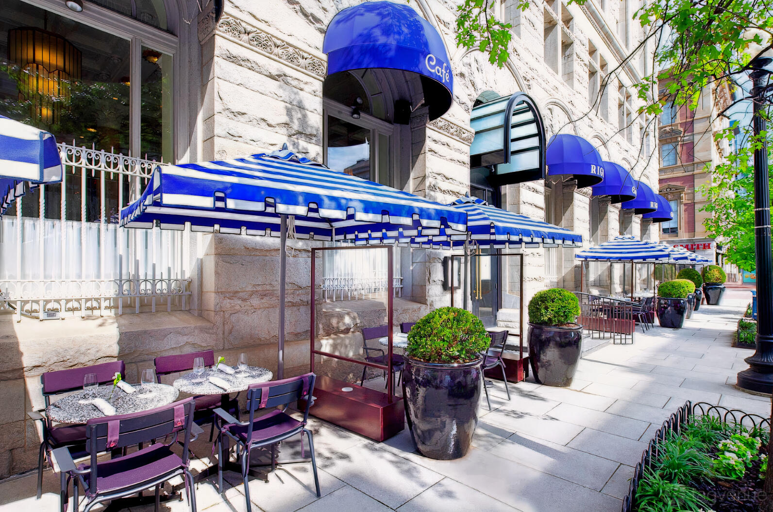 An outdoor restaurant patio with blue-and-white striped umbrellas, potted plants, and sidewalk seating at Cafe Riggs Board Room.