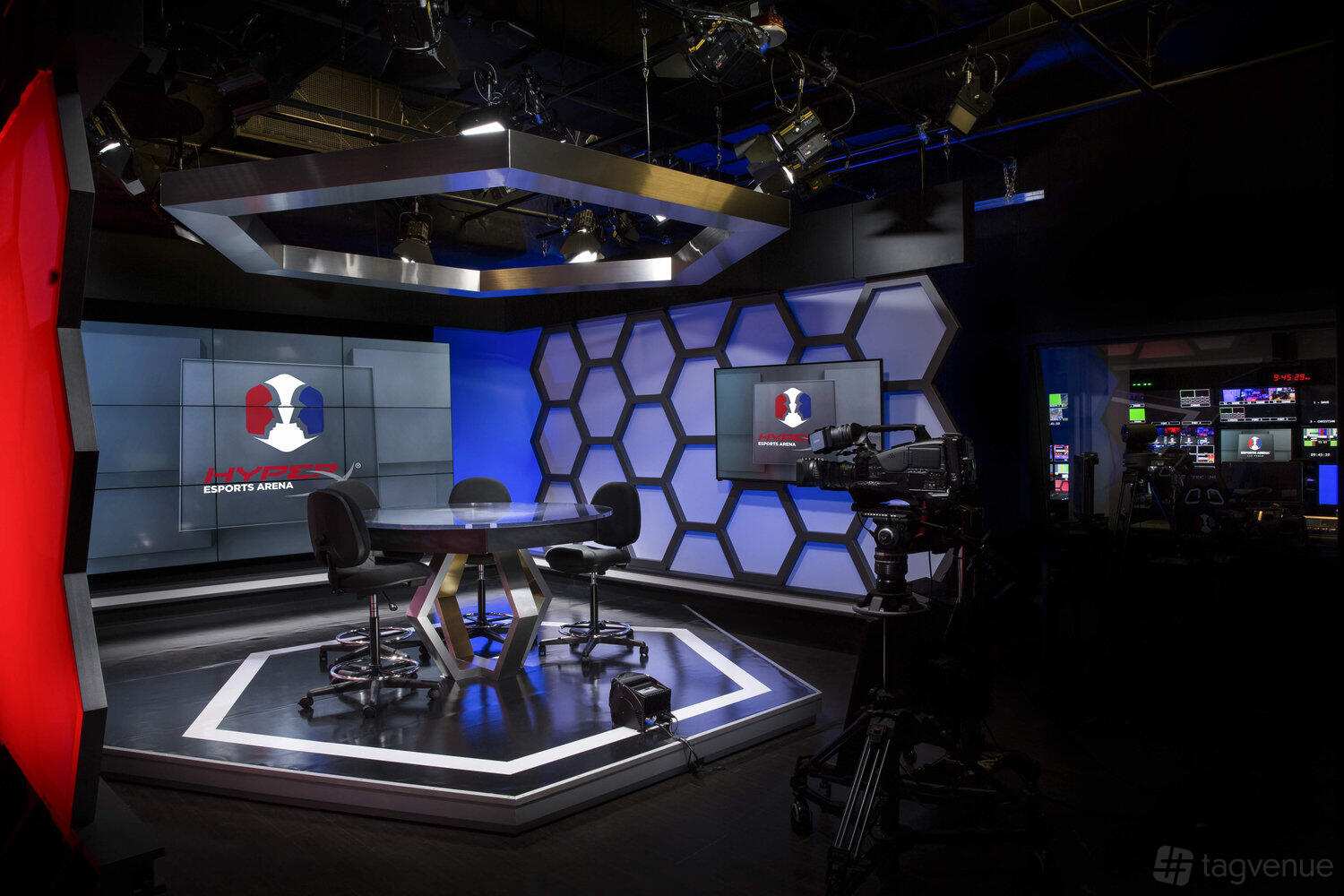 A film studio with illuminated hexagonal wall panels, broadcast cameras, and a modern anchor desk at HyperX Arena Las Vegas.