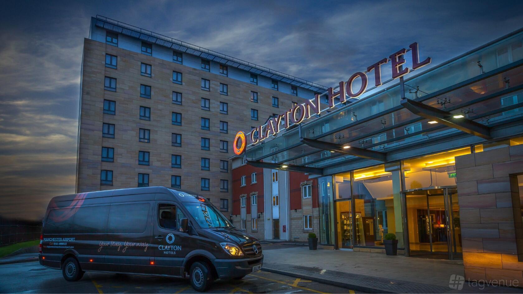 A hotel with a glass-covered entrance, illuminated signage, and parked shuttle van at Clayton Hotel Manchester Airport.
