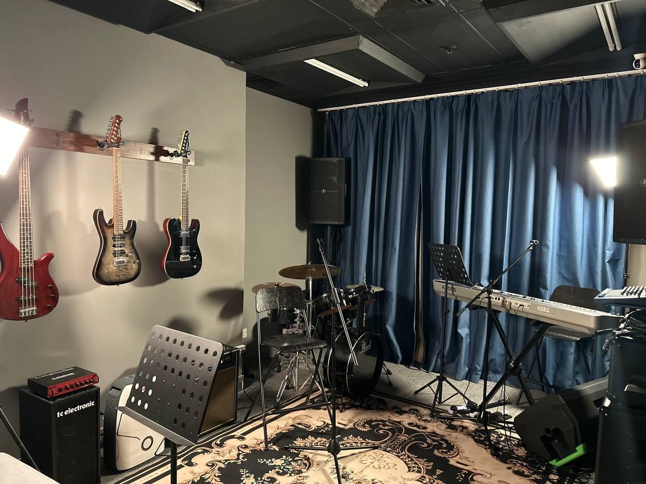A rehearsal room with electric guitars on wall hooks, drum set, keyboard, music stands, and blue curtains at Anvil Music Productions.