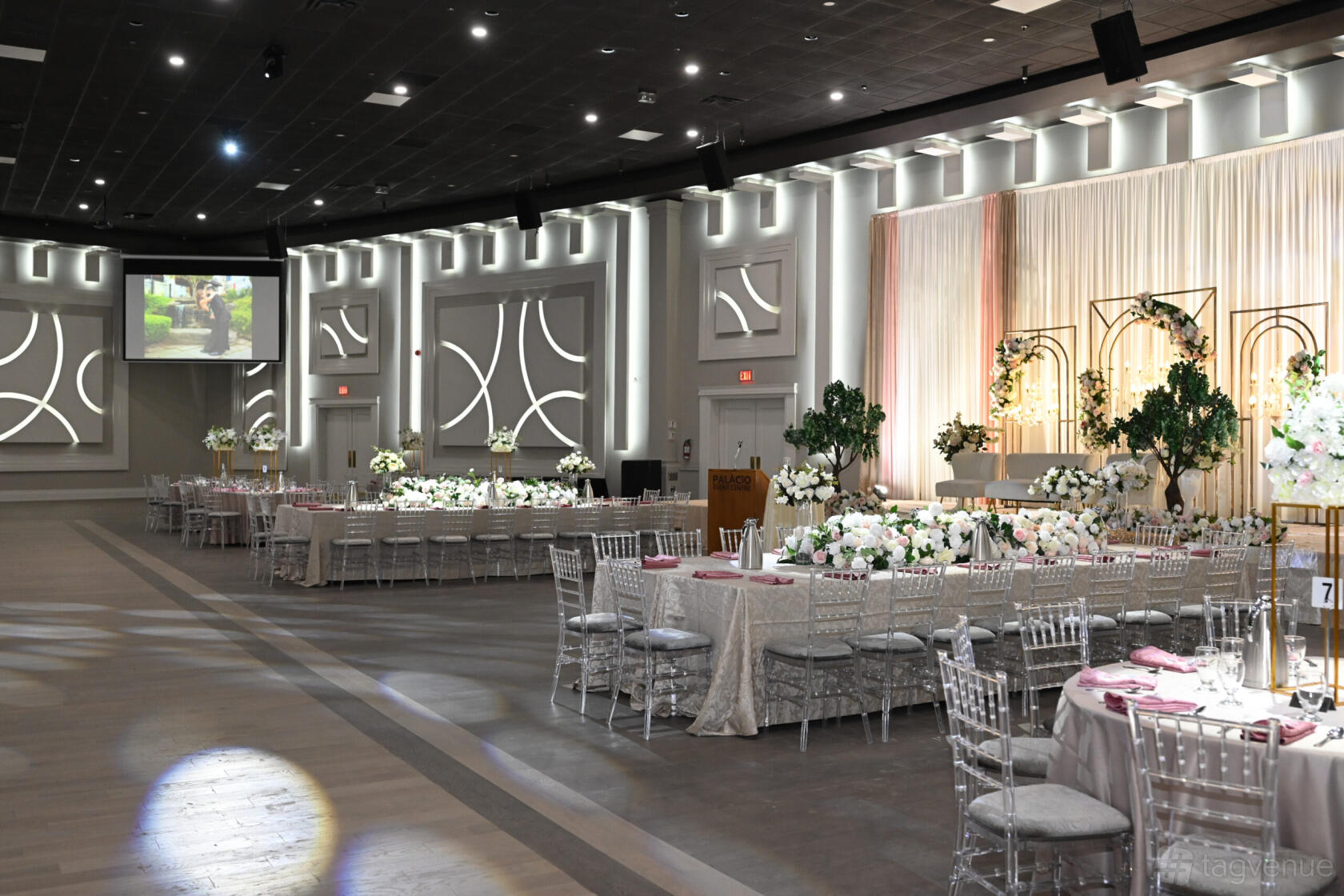 An event venue with draped stage, silver chiavari chairs, long banquet tables, and floral centerpieces at Palacio Event Center.