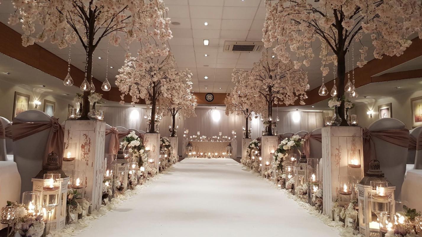 A hotel meeting room with floral archway decor, lanterns, and an aisle lined with pink blossom trees at Cresta Court Hotel.