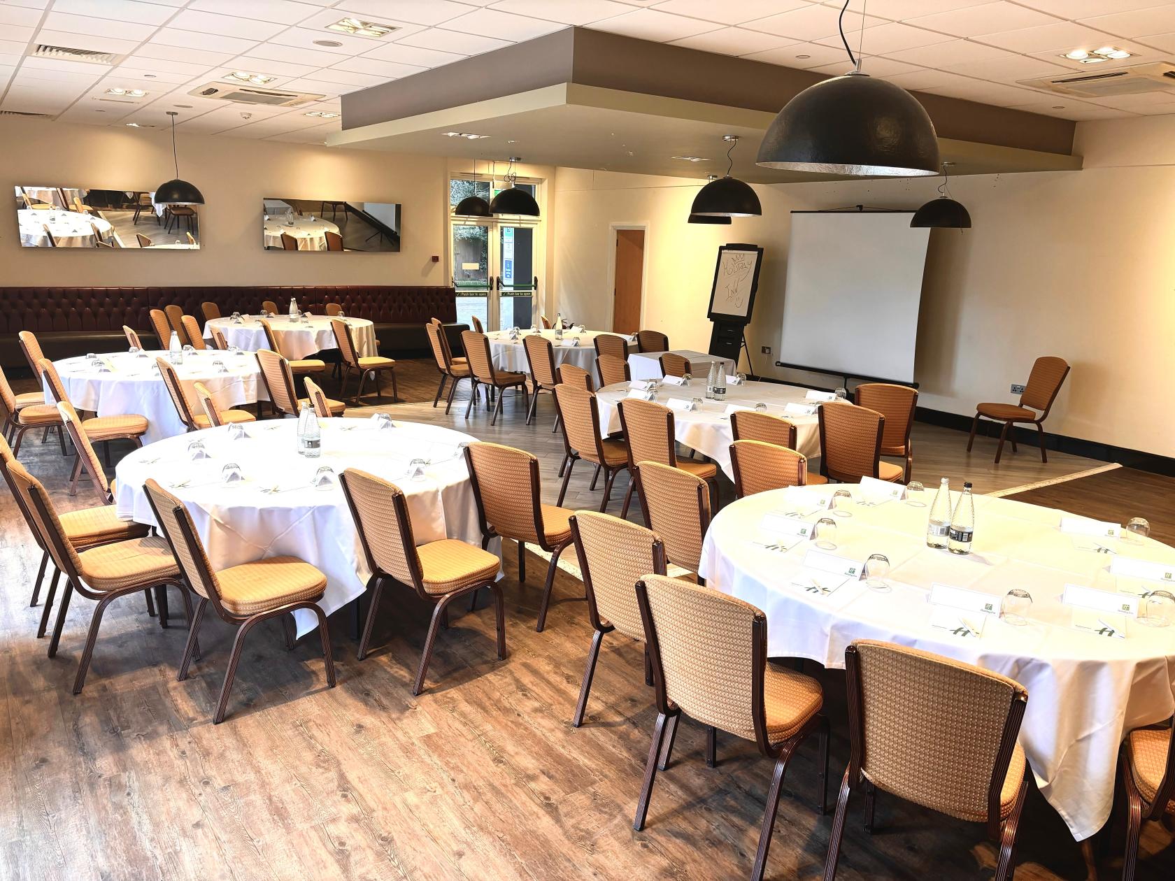 A meeting room with round tables, cushioned chairs, wood flooring, and a projector screen at Holiday Inn Kenilworth-Warwick.