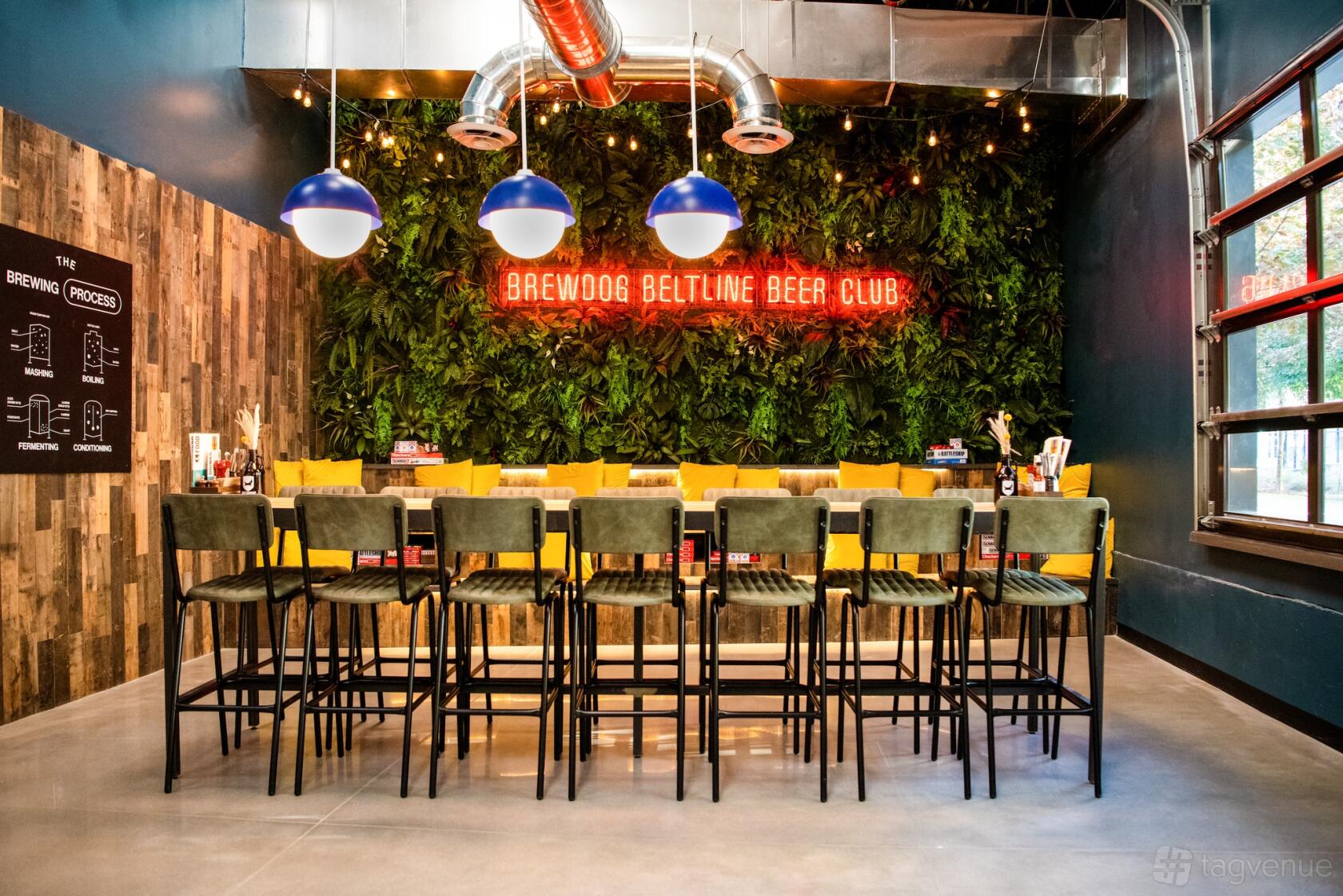A bar with green velvet stools, a living plant wall, exposed ductwork, and neon signage at BrewDog Atlanta.