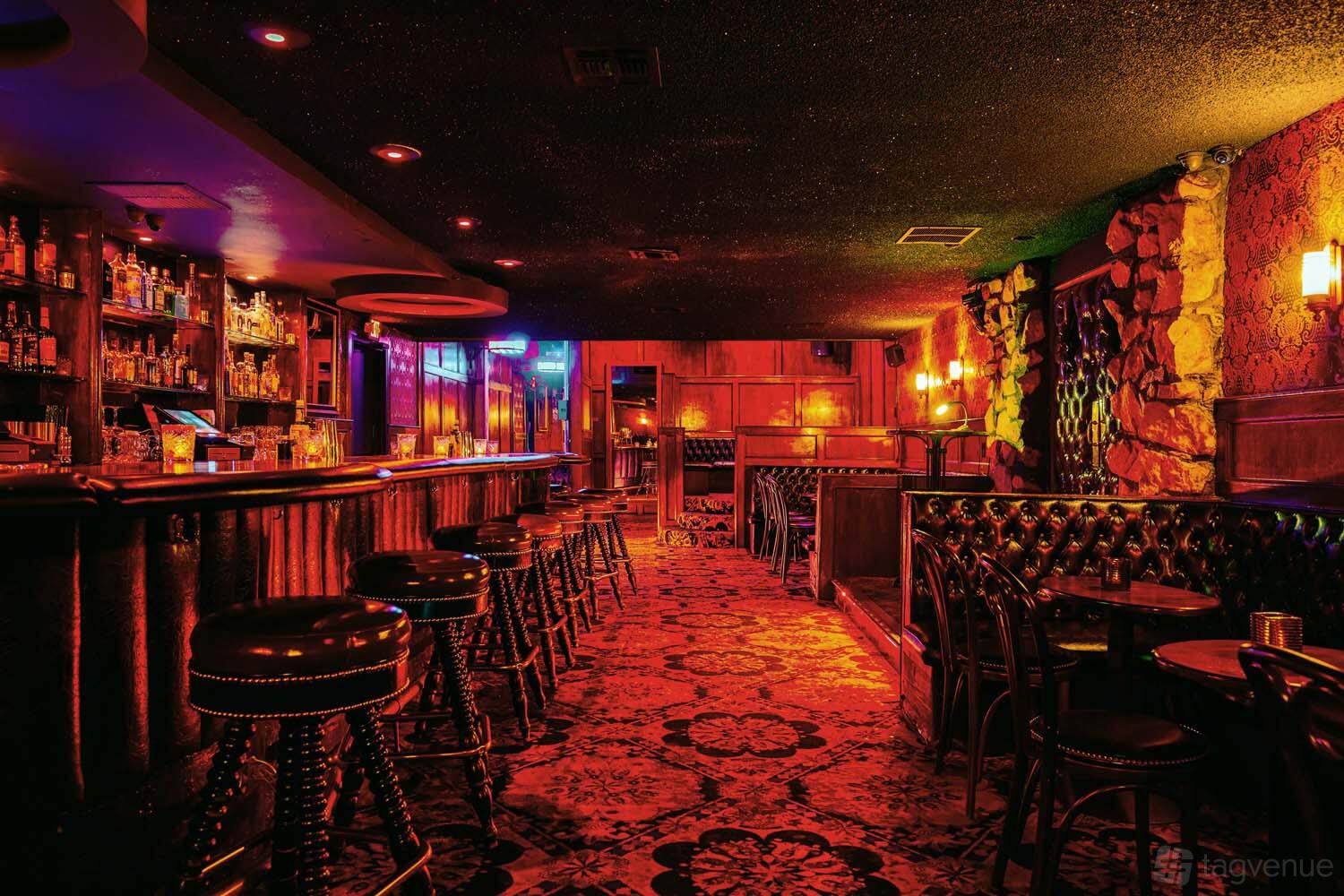 A lounge with patterned carpet, dark wood paneling, leather bar stools, and a stocked bar at The Three Clubs.