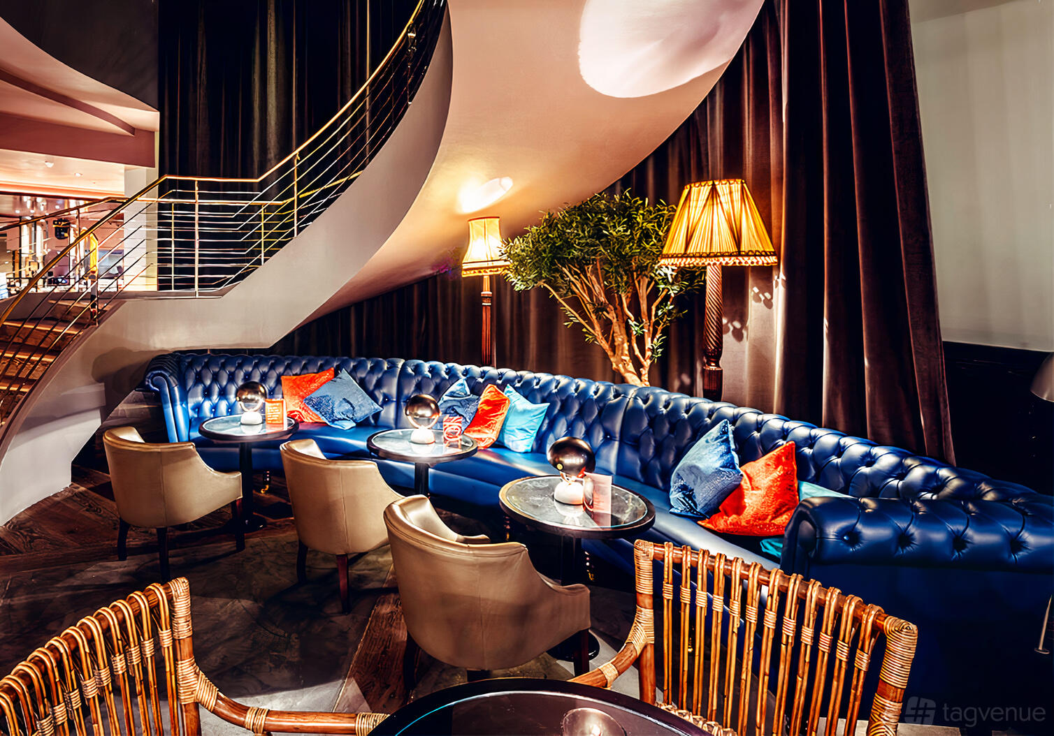 An event space in a restaurant with a blue tufted banquette, round tables, and gold floor lamps at 100 Wardour Street.