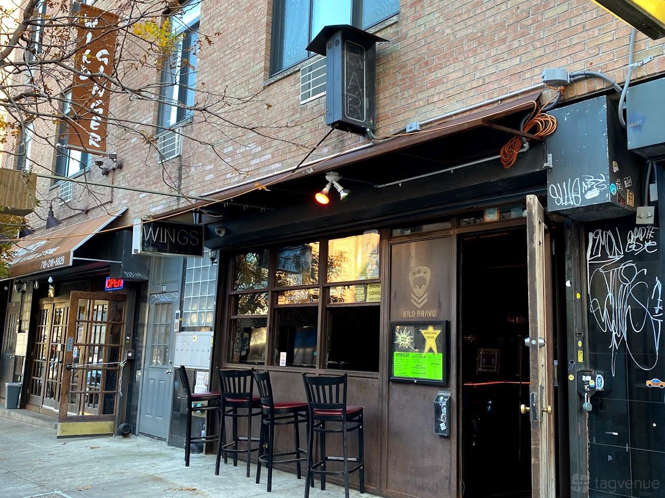 A pub with a brick facade, sidewalk seating with barstools, and open front doors at Kilo Bravo.