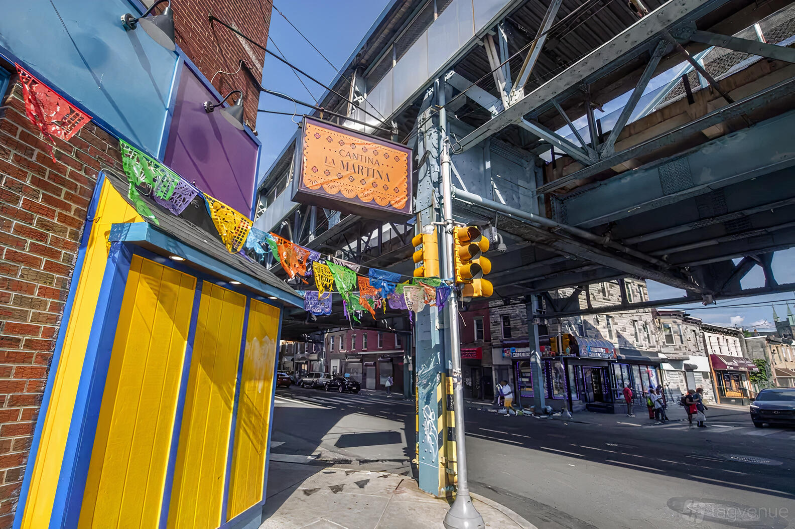 A restaurant with colorful storefront, papel picado banners, and a hanging sign under an elevated train at Cantina la Martina.