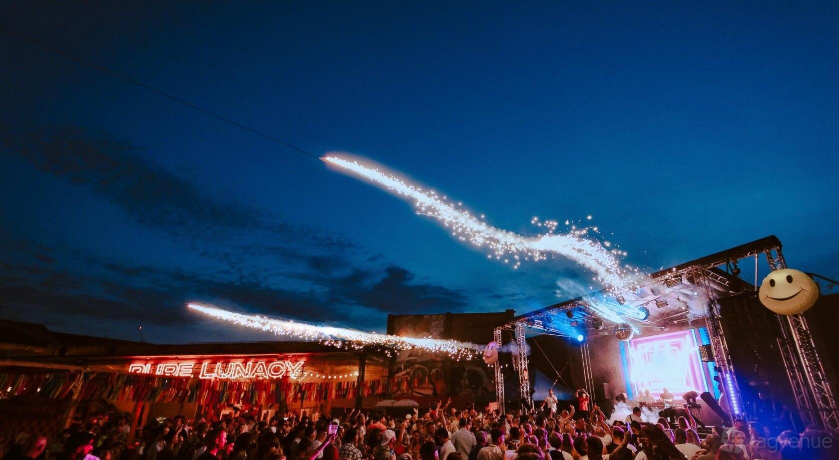 An open air venue with a large crowd, stage lighting, fireworks, and a neon sign at Luna Springs.