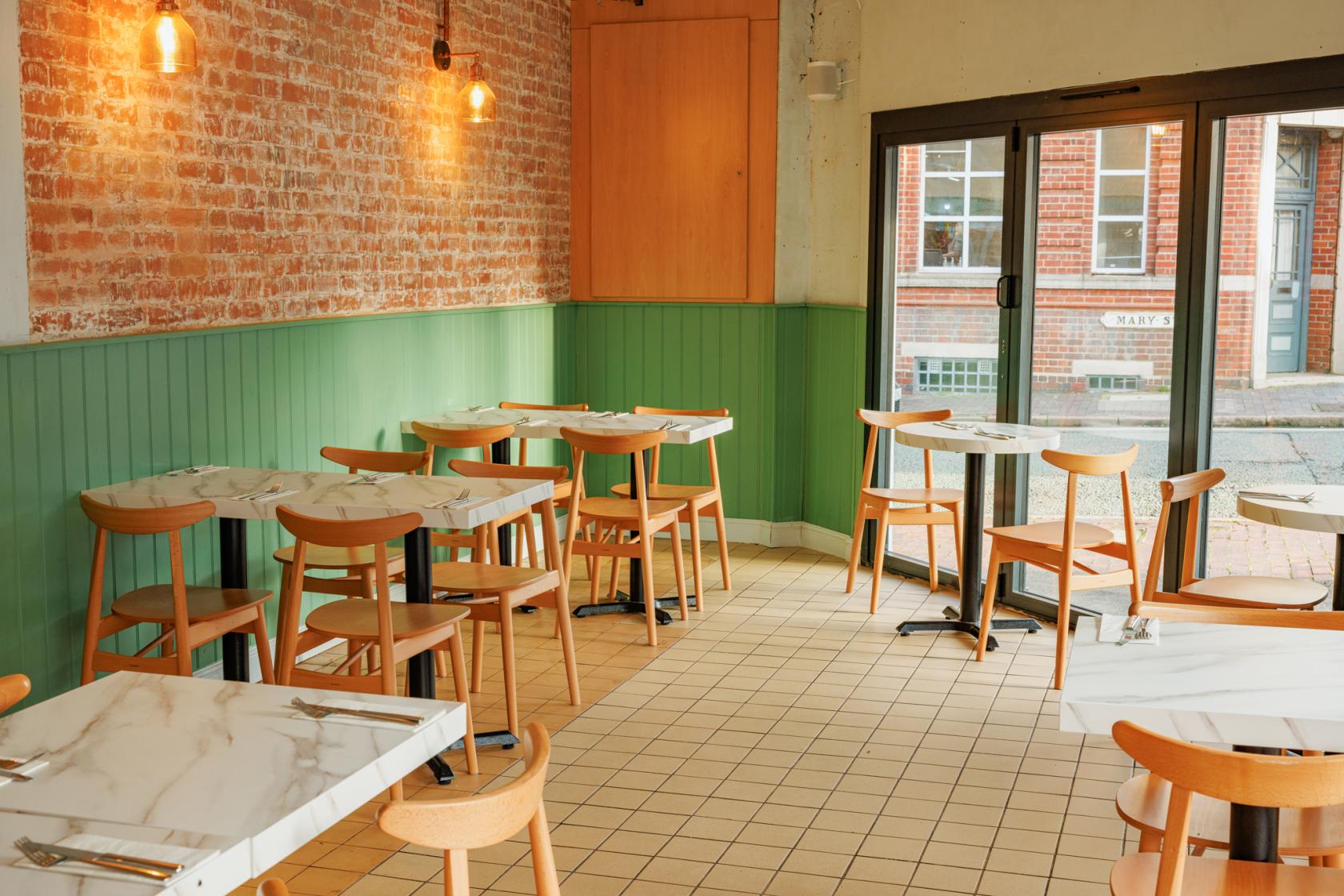 A restaurant dining area with marble-topped tables, wooden chairs, exposed brick walls, and large windows at Trentina.