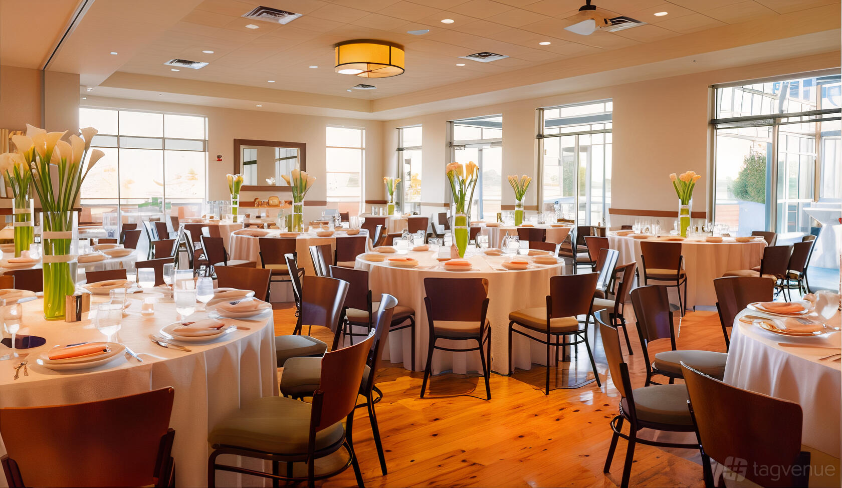 A banquet room with round tables, white linens, tall floral centerpieces, and large windows at Pinstripes - Oakbrook.