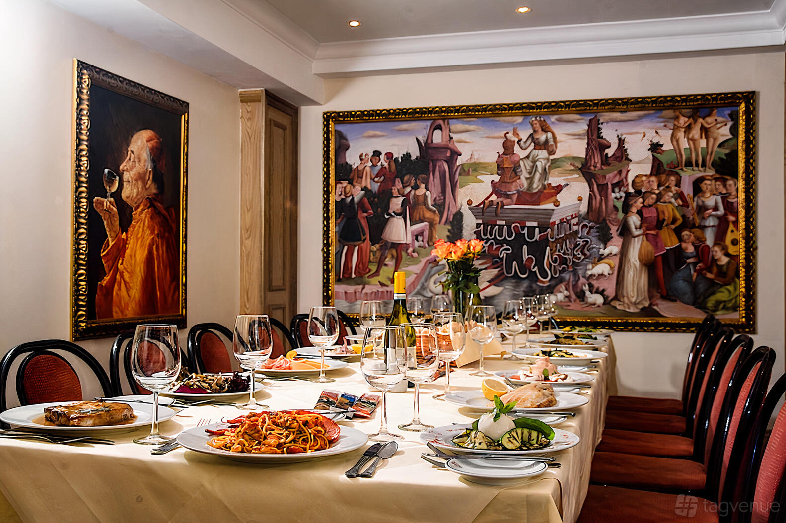 A dining room with a long table set for a meal, surrounded by red chairs and large framed paintings at Bolton's Venetian Room.