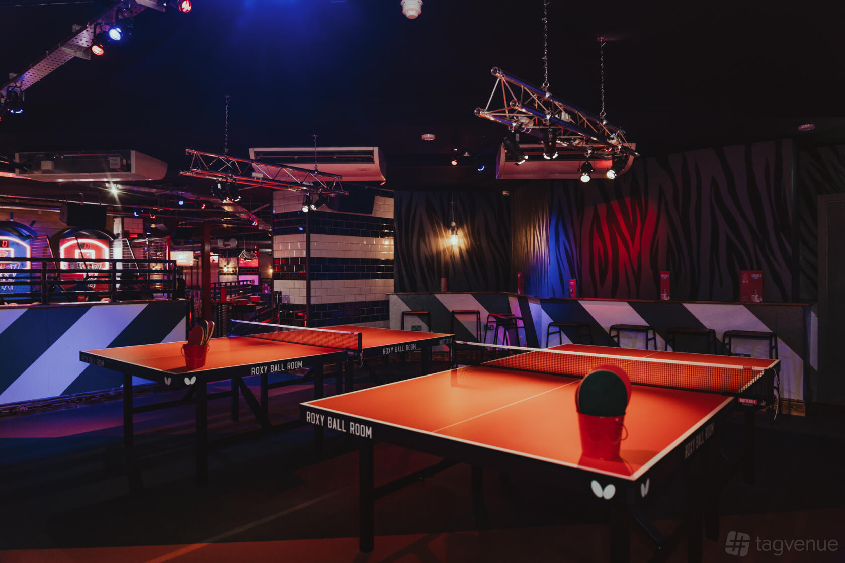 An indoor event space with orange ping pong tables, black stools, and colorful lighting at Roxy Ball Room Victoria Square.