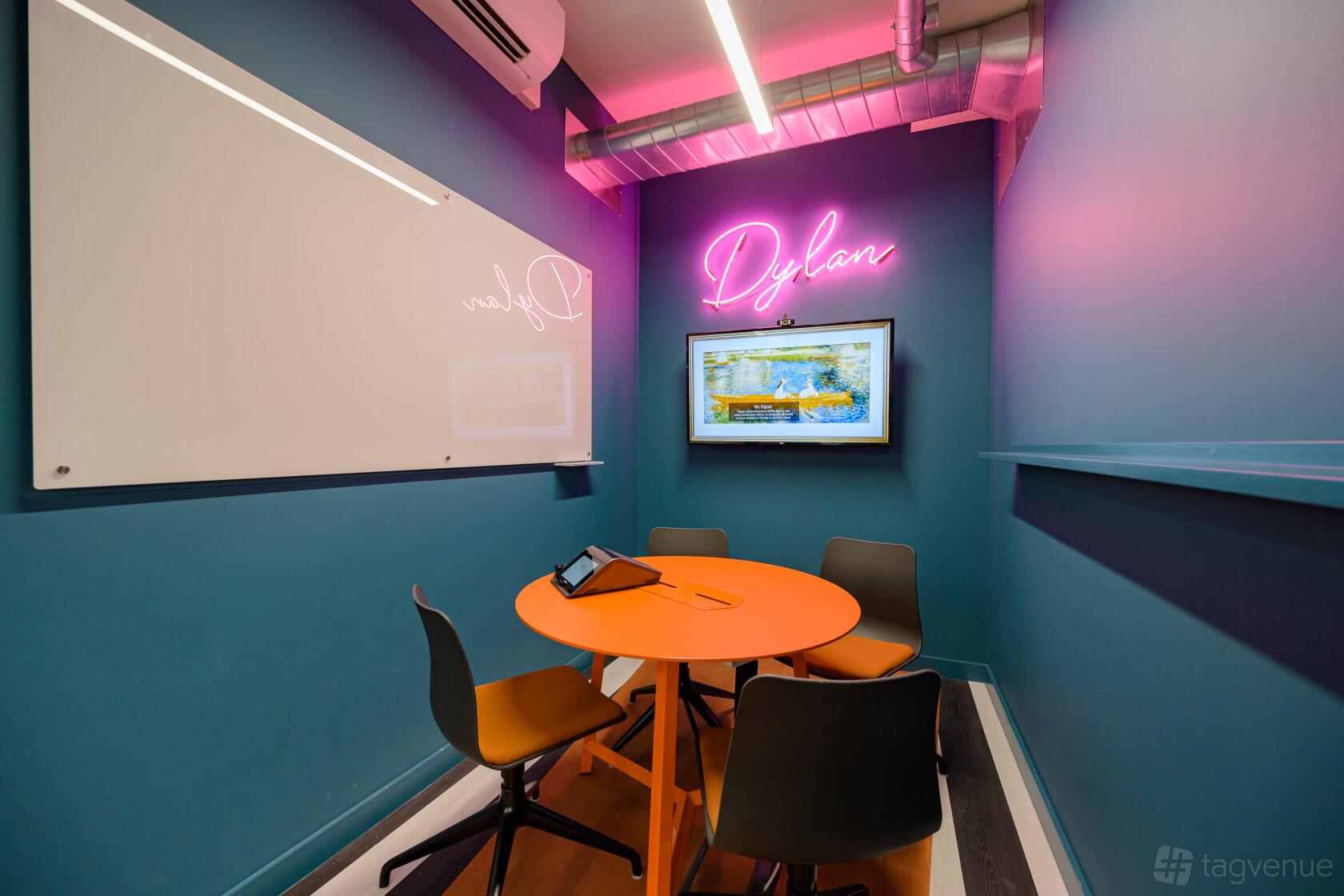 A small meeting room with teal walls, a round orange table, wall-mounted screen, and neon sign at Huckletree Ancoats.