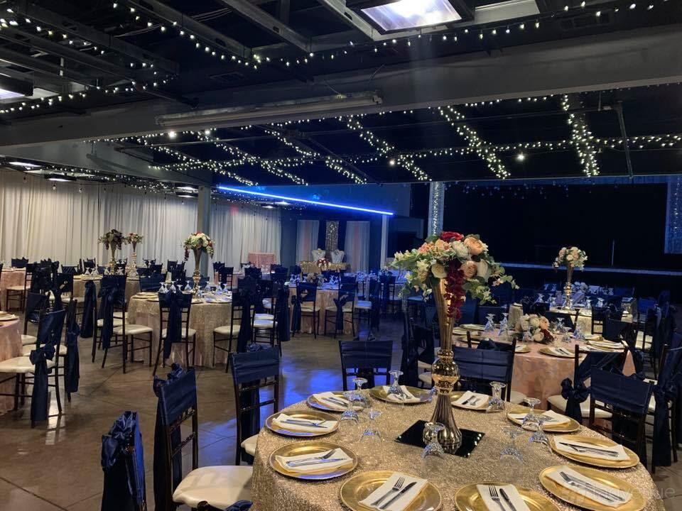 An event hall with round tables, floral centerpieces, navy chairs, and ceiling string lights at Hobby Event Center.