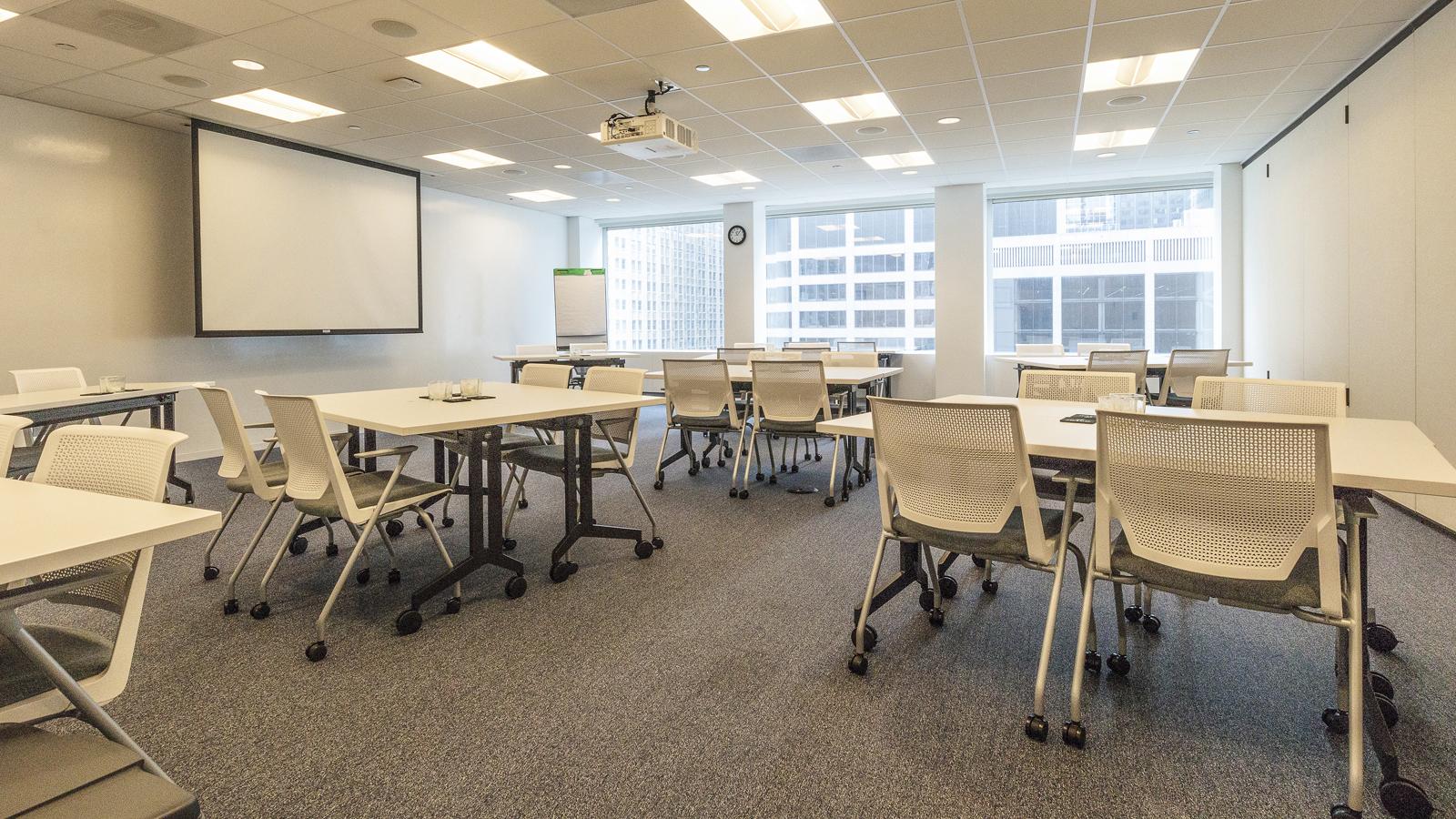 A training room with movable tables, white mesh chairs, a ceiling projector, and large windows at Association Forum.