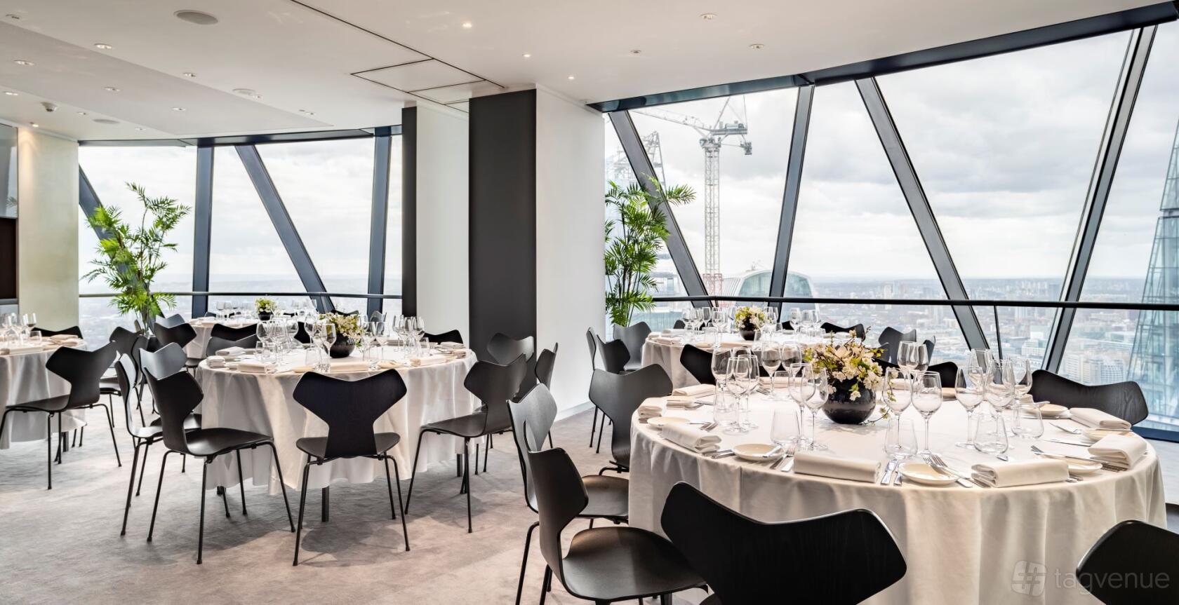 A function room with floor-to-ceiling windows, round tables dressed in white linens, and city skyline views at Searcys at The Gherkin.