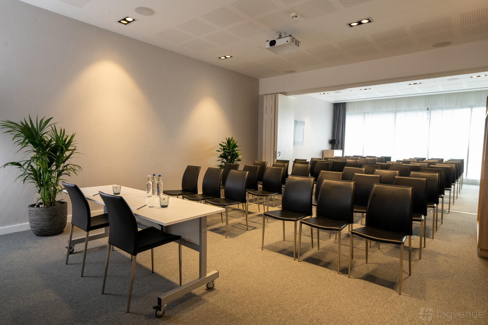 A hotel meeting room with rows of black chairs, a presenter’s table, carpeting, and potted plants at INNSiDE by Melia Liverpool.