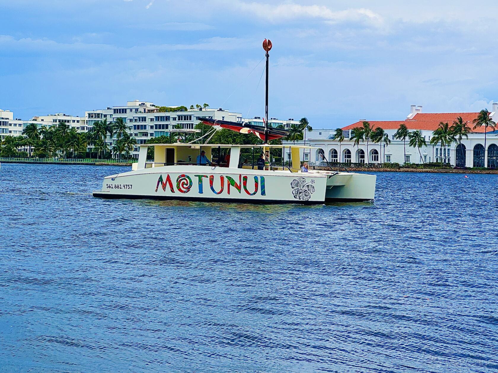 A yacht with a spacious deck and colorful Montunui lettering cruising near waterfront buildings at Lots Of Yachts/Lots Of Spots South Florida.
