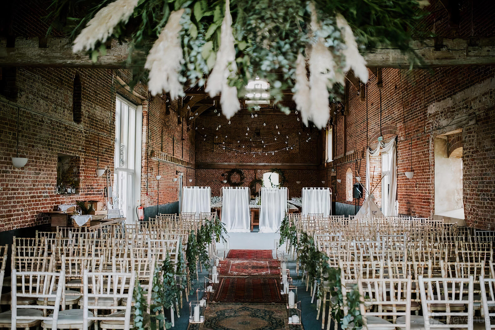 A barn with exposed brick walls, high beamed ceilings, string lights, and rows of wooden chairs at Godwick Great Barn.