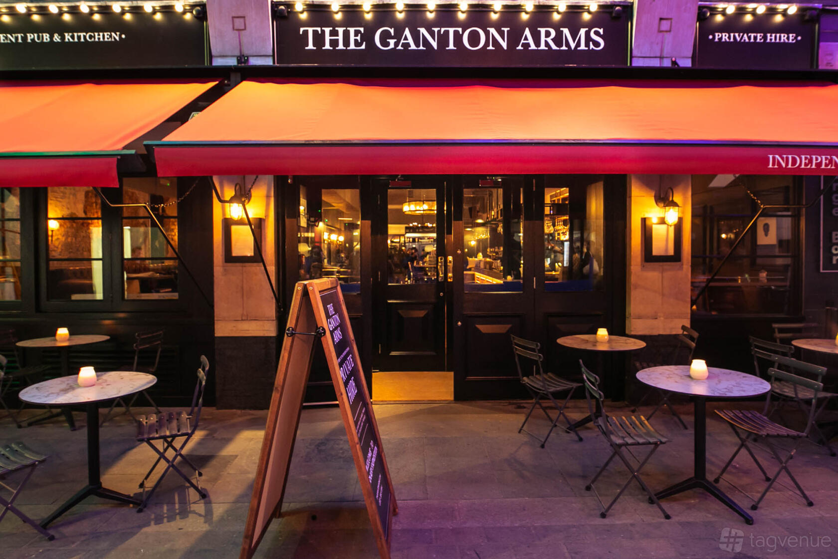 A pub with outdoor bistro tables, folding chairs, and red awnings at The Ganton Arms.