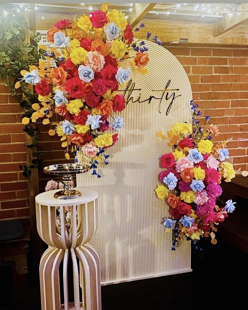 An event space in a bar with a flower wall display, colorful artificial flowers, and exposed brick at The Purple Emerald Lounge Bar.