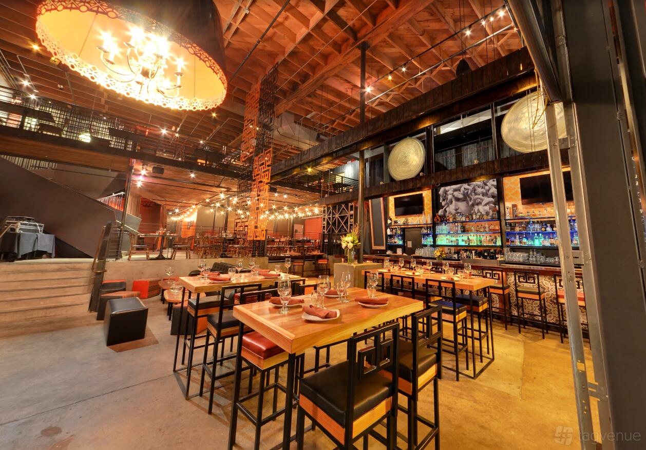 A restaurant with high exposed wood ceilings, hanging string lights, and a colorful backlit bar at Meze Greek Fusion.