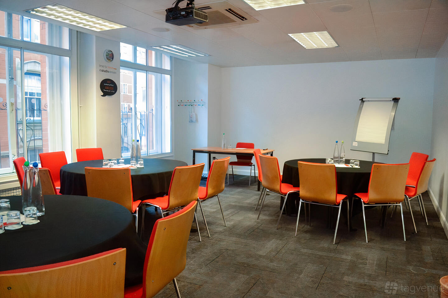 A meeting room with round tables, red chairs, large windows, and a flipchart stand at The Studio Birmingham.