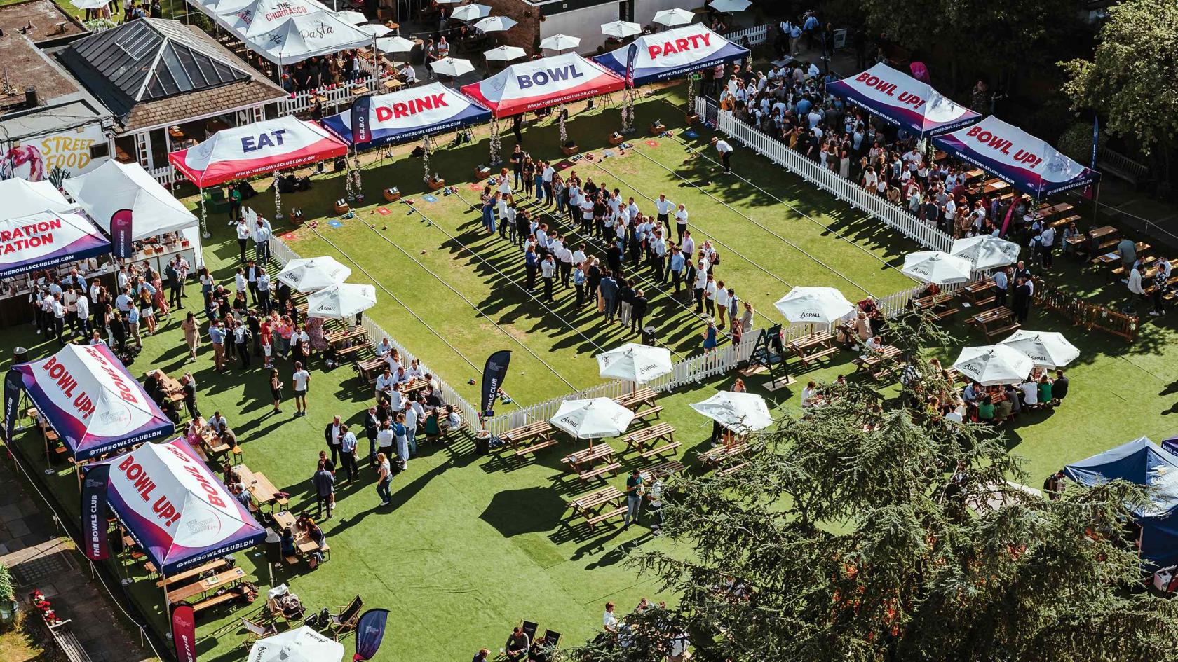 An outdoor event venue with lawn bowling greens, picnic benches, and canopy tents at The Bowls Club.