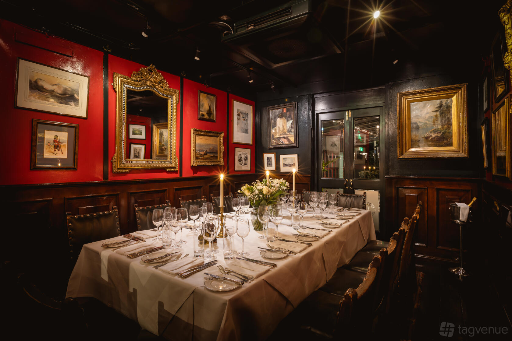 A restaurant dining room with dark wood paneling, red walls, framed artwork, and a long table set with candles at Boisdale of Belgravia.