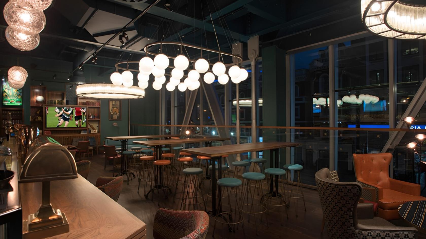 A lounge in a pub with globe pendant lights, high tables, bar stools, and floor-to-ceiling windows at Greenwood.