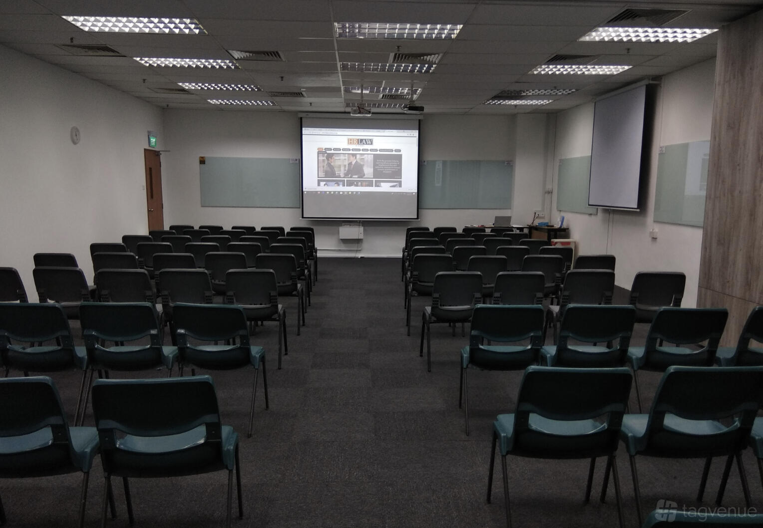 A training room with rows of black chairs, a projector screen, and whiteboards at Synergy Room Singapore Shopping Centre.