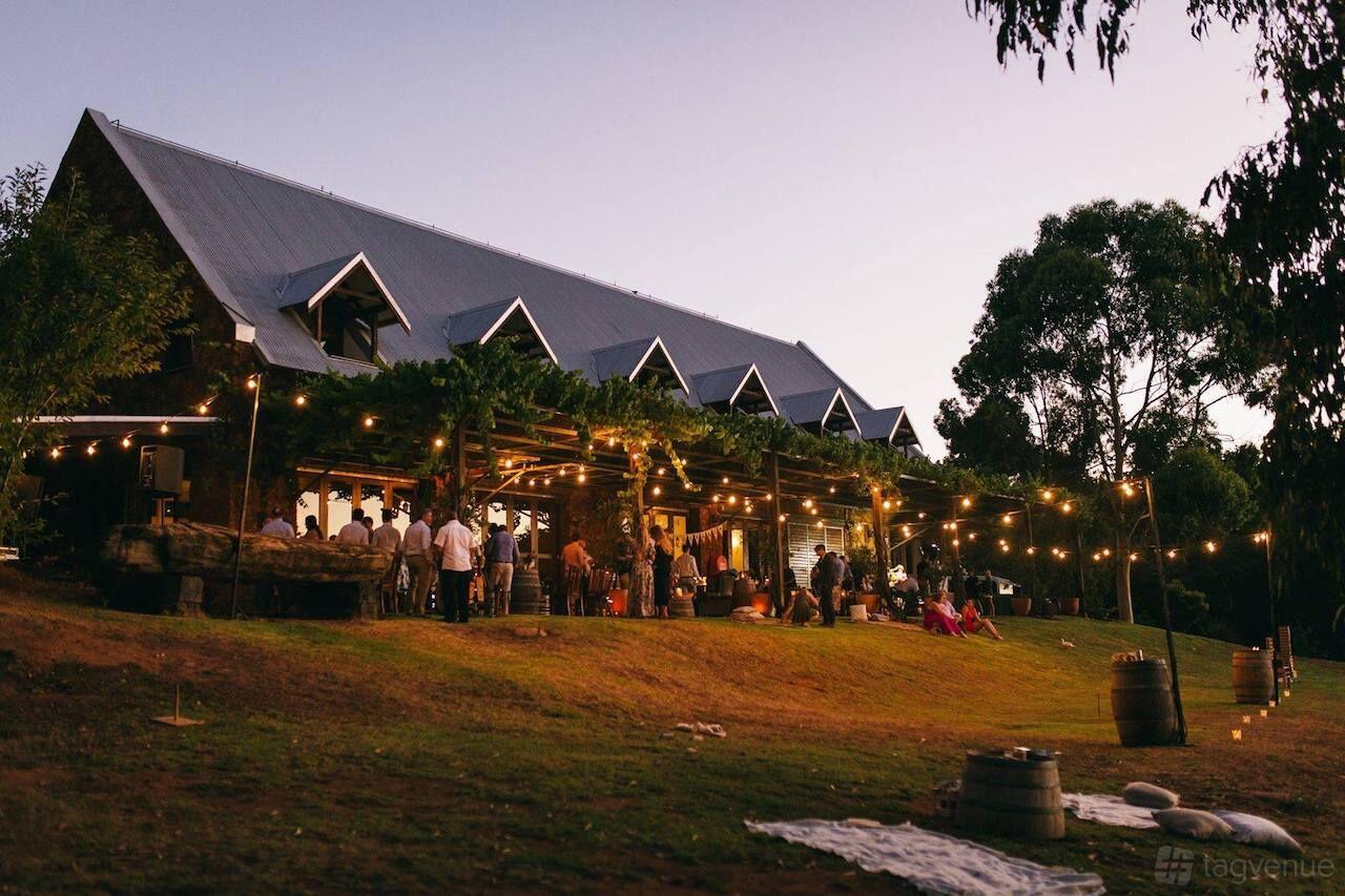 A rustic event venue with string lights, exposed timber beams, and a lawn area at Stonebarn Weddings.