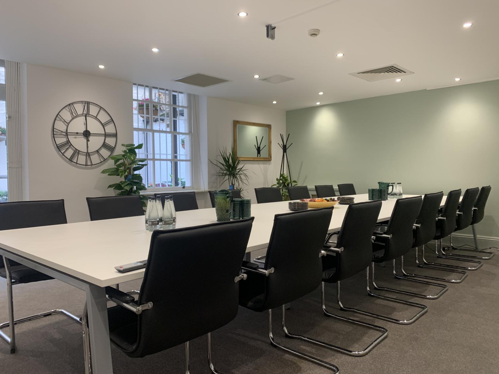 A meeting room with a long white table, black chairs, potted plants, wall clock, and large windows at Citibase London Victoria Catherine Place.