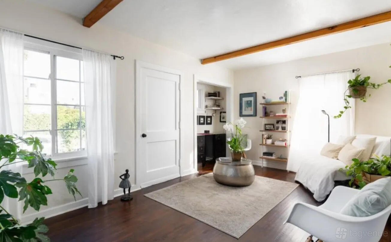 A living room with hardwood floors, large windows, potted plants, and a neutral area rug at Bohemian House.