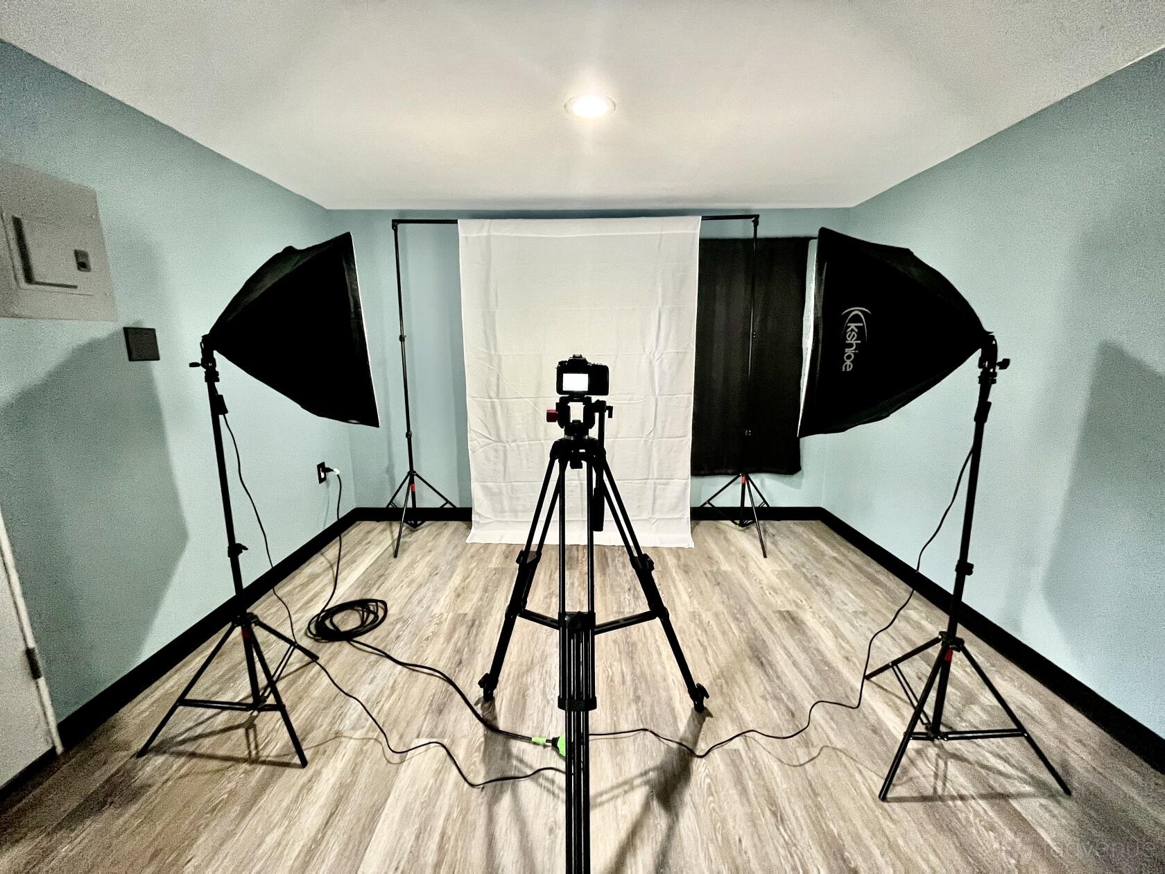 A photo studio with a white backdrop, two softbox lights, and a camera on a tripod at Revolving Red Productions.