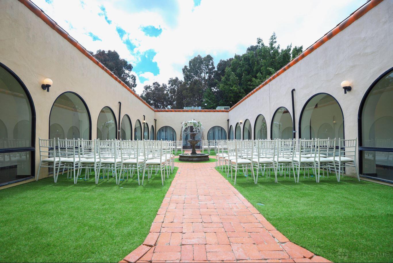 An event venue patio with white chairs arranged on artificial grass, brick walkway, and a central fountain at Spark at Encore Event Center.