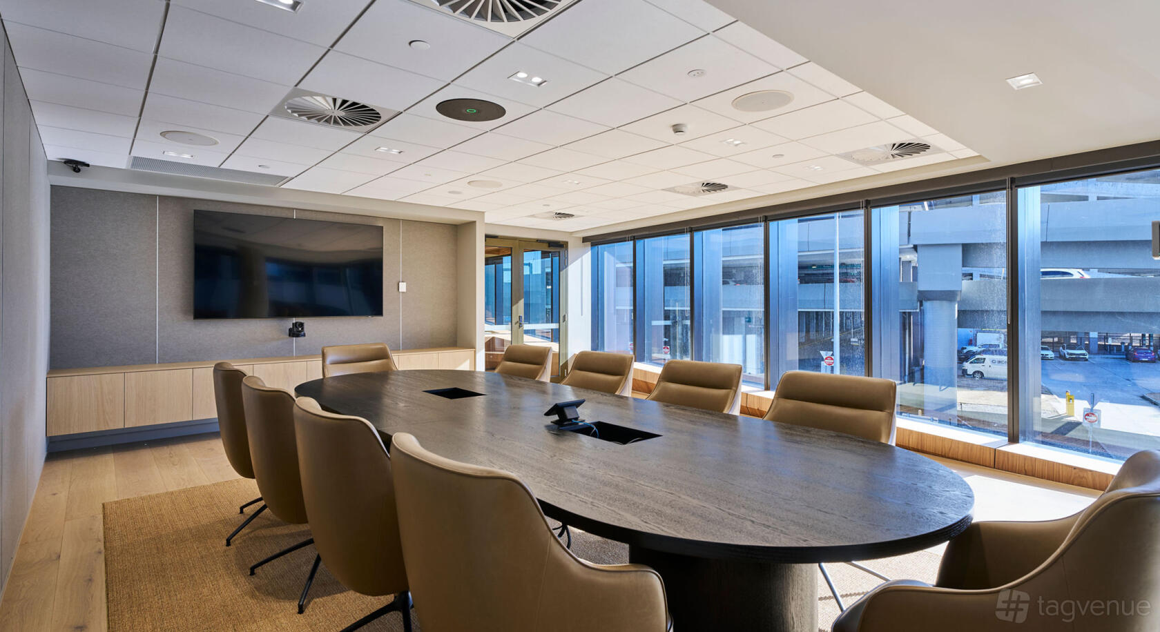 A meeting room with a large oval conference table, leather chairs, wall-mounted screen, and floor-to-ceiling windows at Novotel Melbourne Airport.