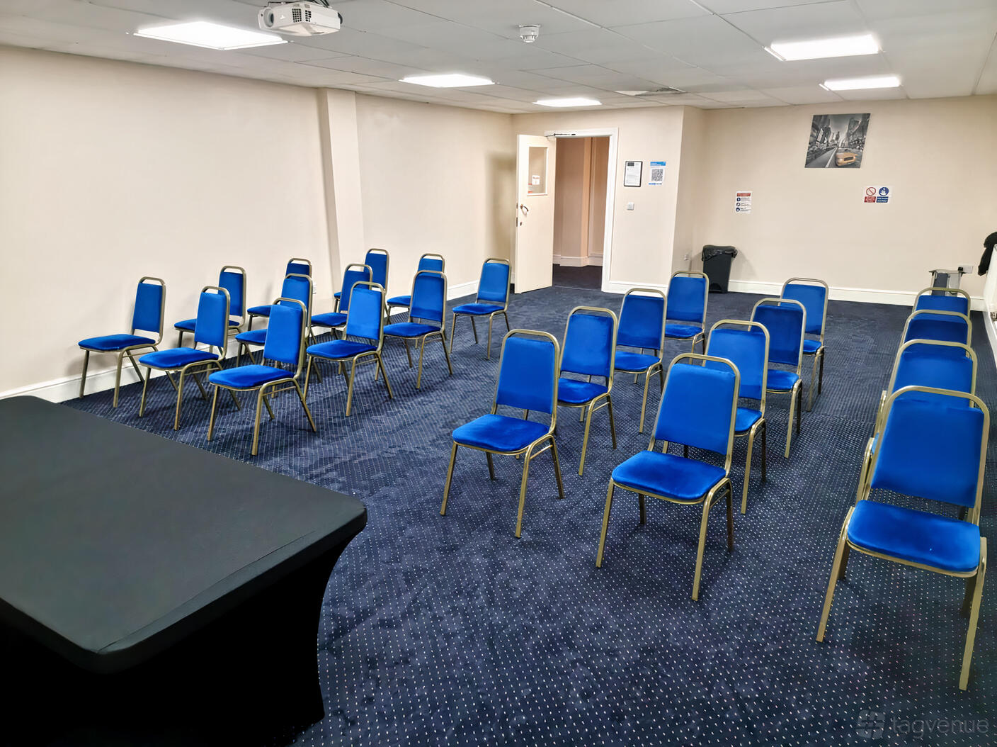 A meeting room with rows of blue chairs on a navy carpet and a black-clothed table at The Saffron Centre.