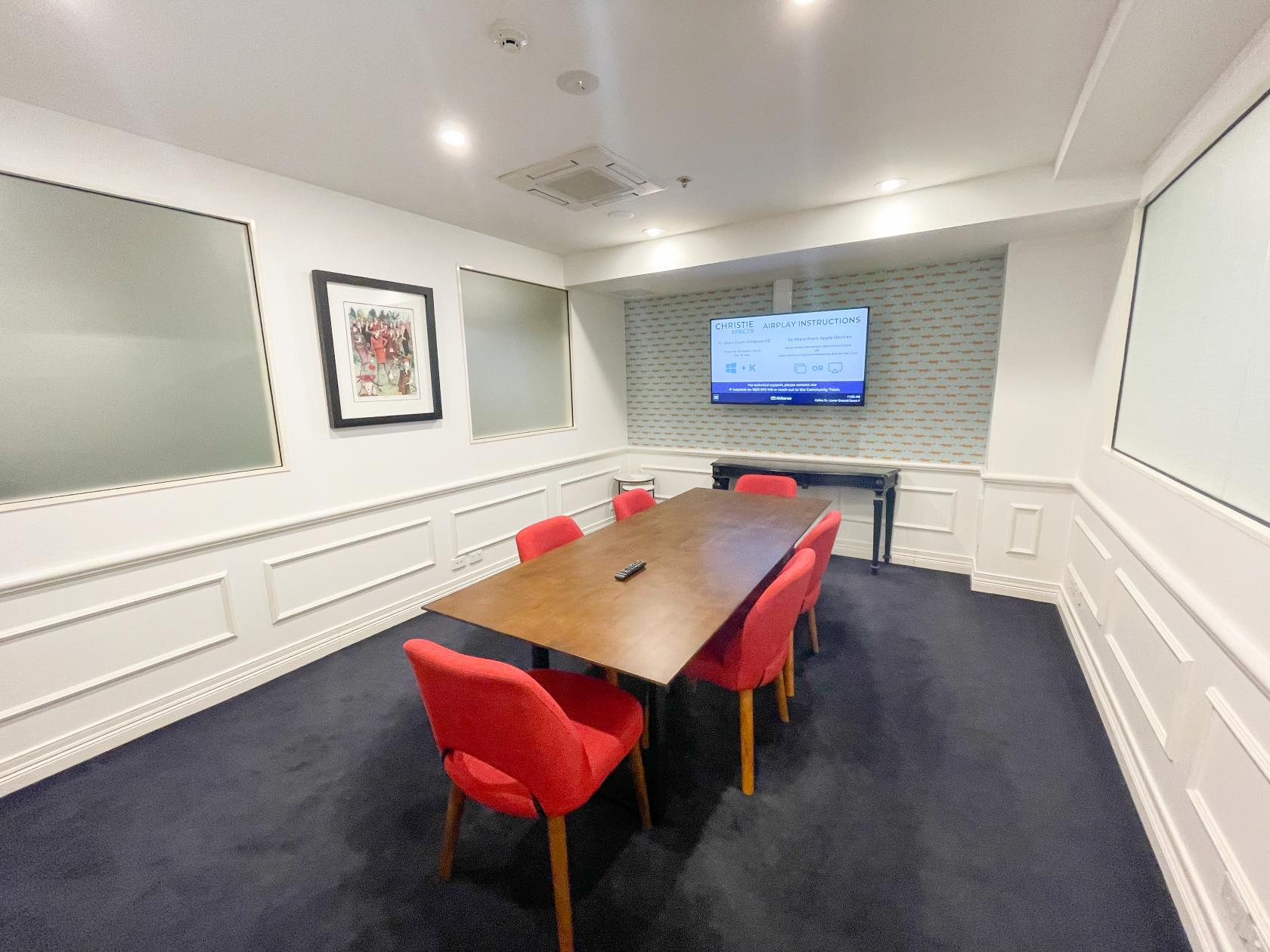 A meeting room with a rectangular wooden table, red chairs, wall-mounted screen, and framed artwork at Christie Spaces Conferencing Melbourne.