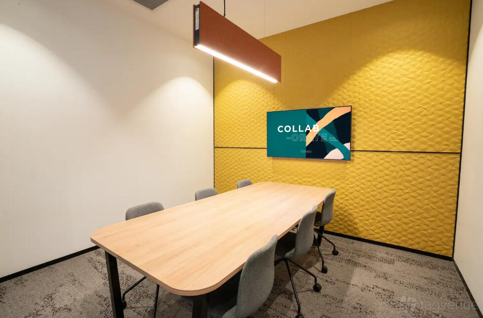 A coworking meeting room with a long wooden table, gray chairs, yellow textured wall, and wall-mounted screen at Waterman at Eastland.