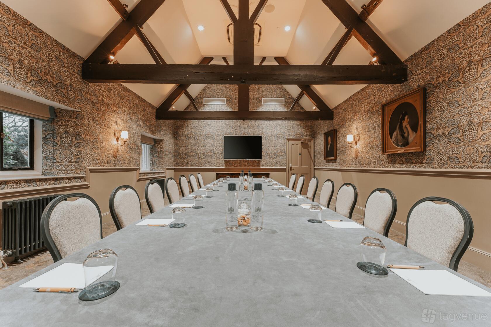 A meeting room with a long conference table, vaulted timber beams, patterned wallpaper, and a wall-mounted TV at Ellenborough Park.