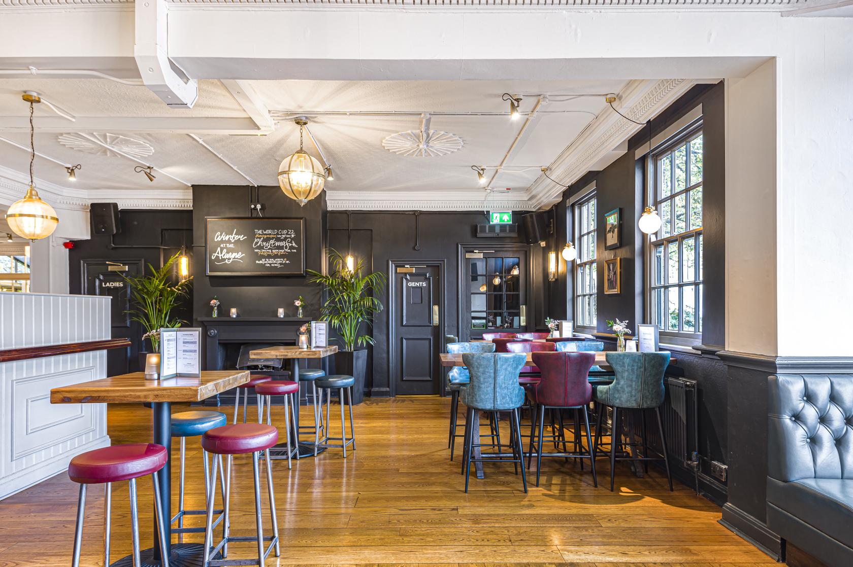 A pub with wooden floors, dark walls, large windows, velvet chairs, and globe pendant lighting at The Alwyne Castle.