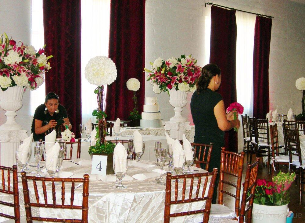 A community centre with round tables draped in white linens, floral centerpieces, and tall windows with burgundy curtains at Woman's Club of Hollywood.