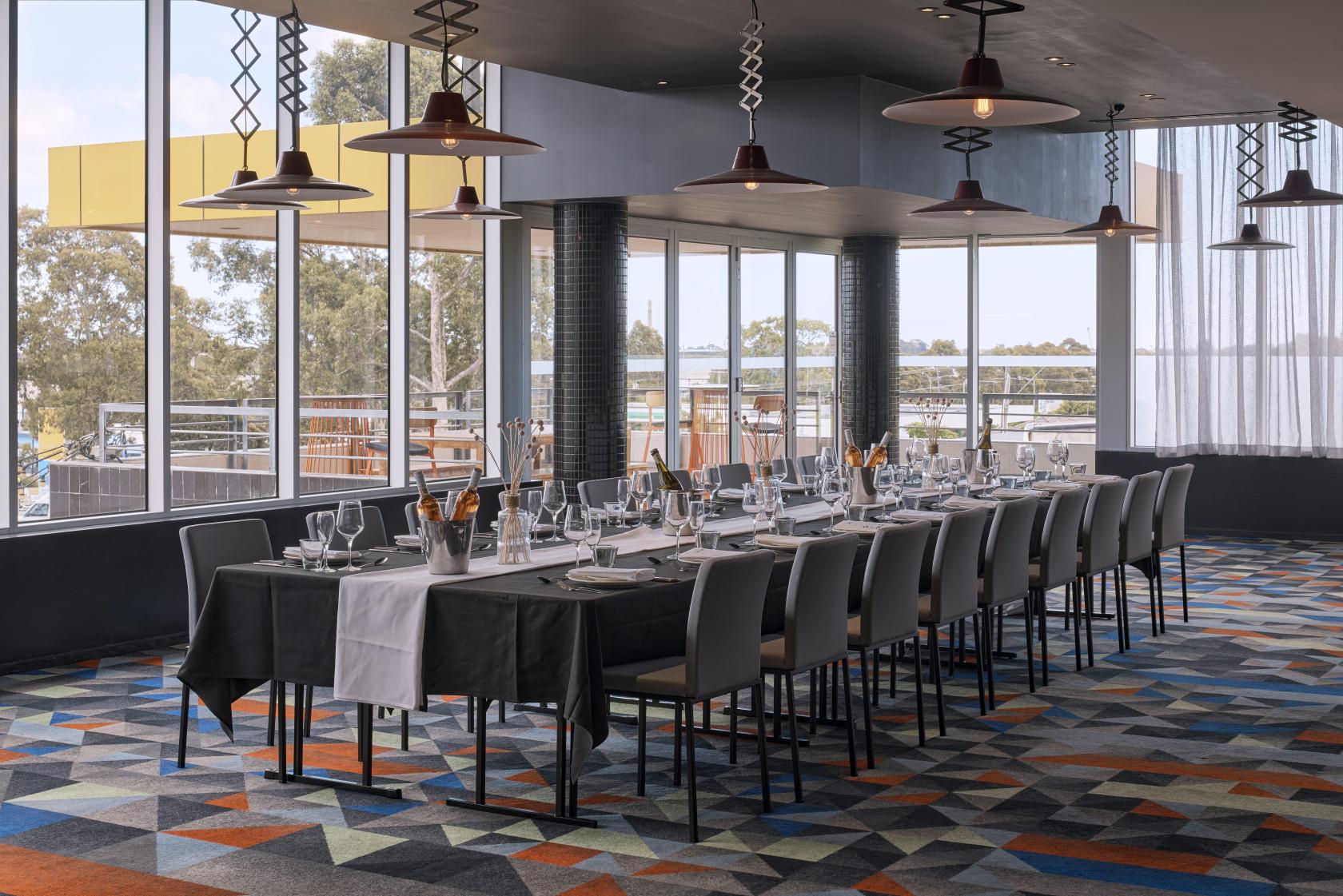 A restaurant with floor-to-ceiling windows, geometric patterned carpet, and a long table set for dining at Atura Dandenong.