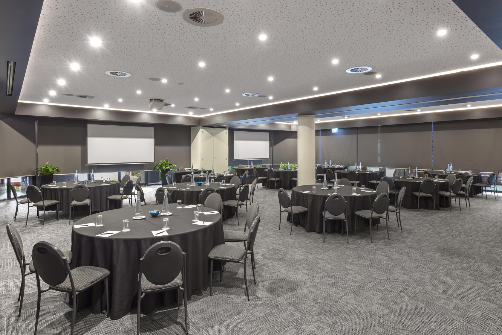 An event space with round tables, grey chairs, two large projection screens, and carpeted floors at Amora Hotel Riverwalk Melbourne.