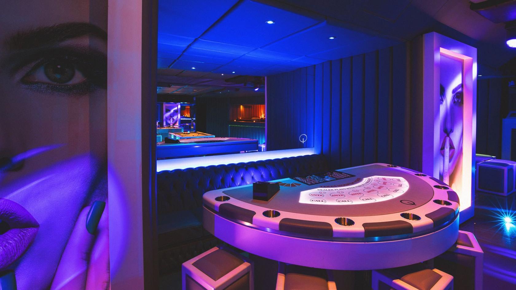 An underground bar space with a poker table, tufted booth seating, blue mood lighting, and graphic wall art at Q Shoreditch.