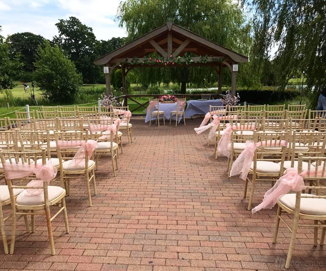 An outdoor event venue with a wooden pergola, rows of chairs draped in pink fabric, and brick paving at Reigate Hill.