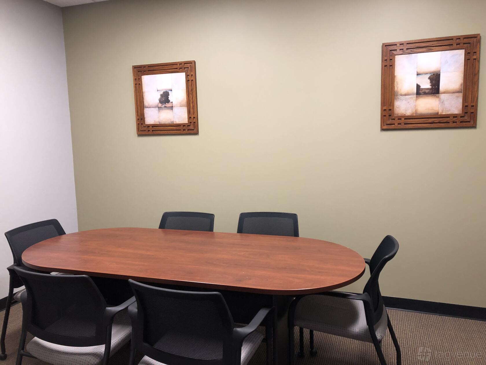 A meeting room with an oval wooden table, eight black chairs, and framed wall art at Workspace@45.